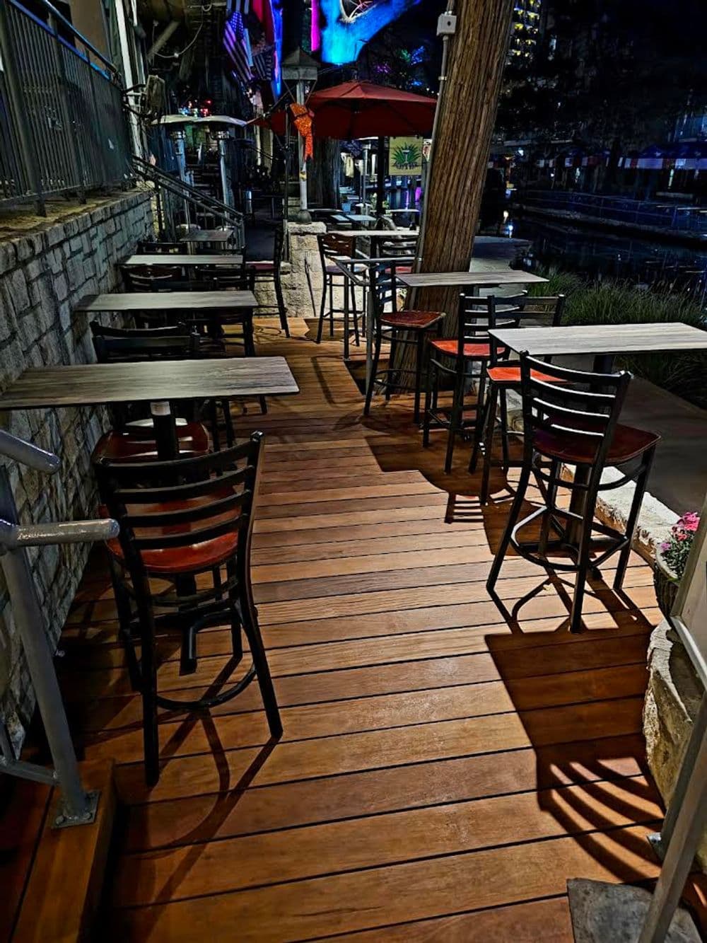 Outdoor seating area with tables and chairs along a wooden deck near a waterway at night.