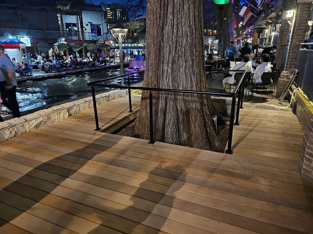 Wooden deck with railing surrounding large tree, overlooking a vibrant riverside dining area.