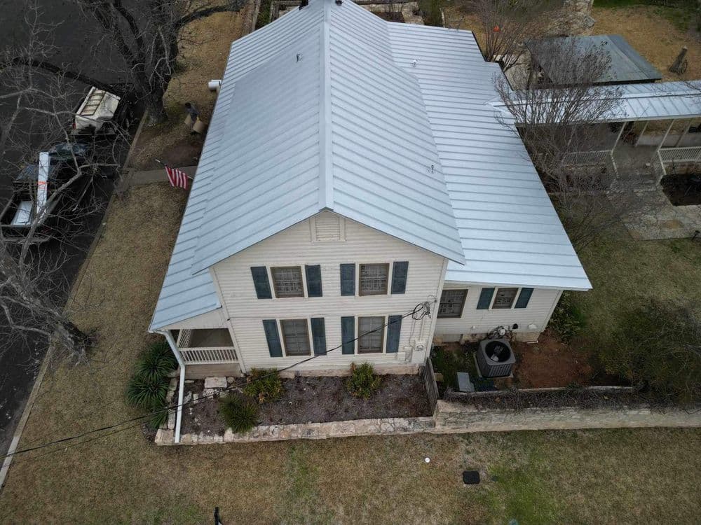 Aerial view of a two-story house with a metal roof, surrounded by trees and grass.