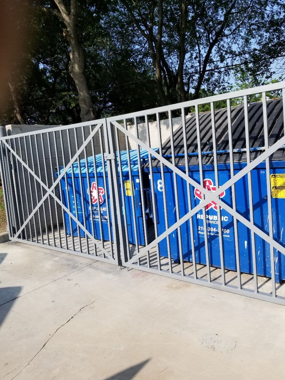 Blue dumpsters behind gated enclosure surrounded by trees in a clean environment.