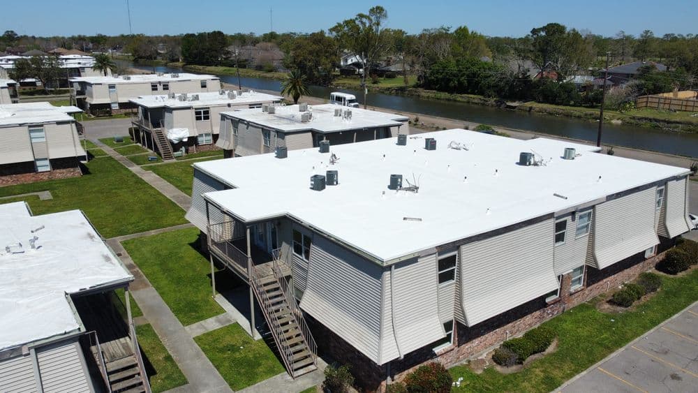 Aerial view of residential apartments with white roofs by a canal, surrounded by green lawns.