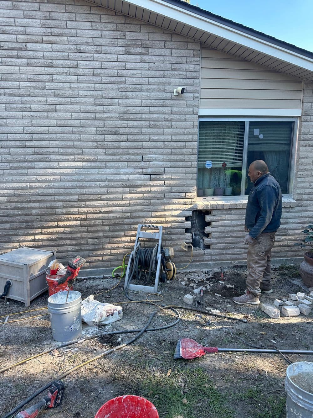 Worker repairs wall with bricks and tools near residential home exterior.