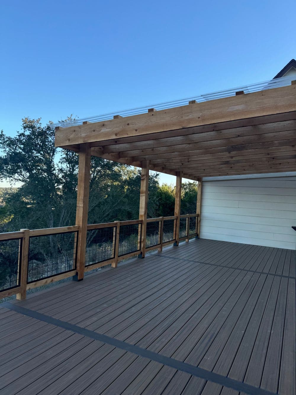 Modern wooden deck with railing under a pergola, featuring clear skies and surrounding trees.