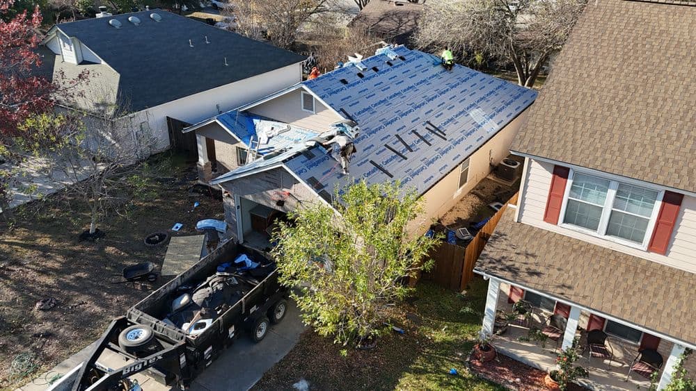 Drone view of a roofing project with workers on a house, surrounded by trees and vehicles.