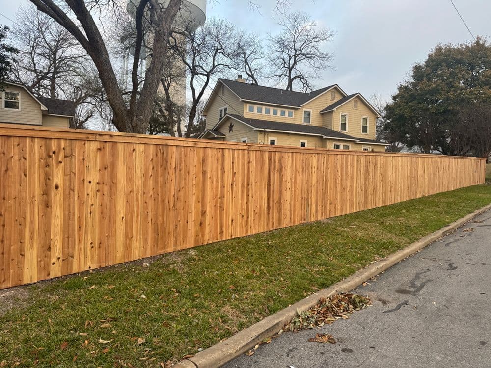 Wooden privacy fence along a residential street with houses and trees in the background.