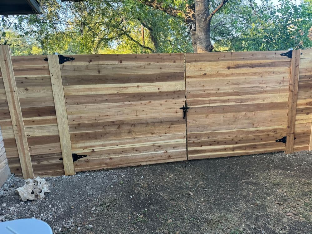 Wooden privacy fence with double gates and black metal hardware in a backyard setting.