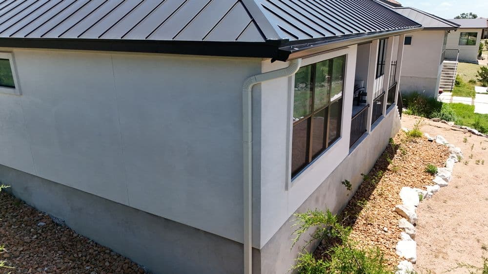 Modern house exterior with metal roof and large windows, surrounded by gravel landscaping.